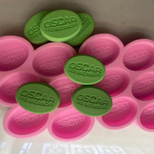 Puede incluir: Un molde de silicona rosa con cavidades ovaladas, que contiene jabones ovalados verdes. Los jabones est&aacute;n estampados con el texto "OSCAR LES VACANCES". El molde es para hacer jab&oacute;n.