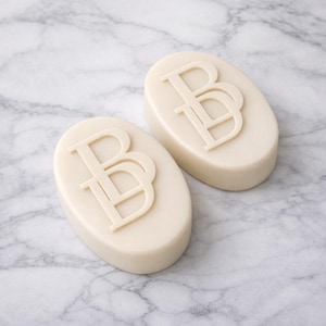 Puede incluir: Dos jabones ovalados de color blanquecino con un monograma "BB" en relieve. Los jabones est&aacute;n colocados sobre un fondo de m&aacute;rmol, creando una est&eacute;tica limpia y elegante. El dise&ntilde;o es simple y moderno, adecuado para diversos usos.