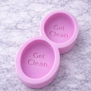 Puede incluir: Un molde de silicona de color lila con dos cavidades ovaladas conectadas. Cada cavidad tiene las palabras "Get Clean" en relieve en el centro. El molde est&aacute; dise&ntilde;ado para hacer jab&oacute;n o manualidades similares.