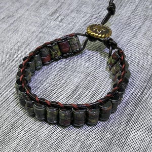 May include: A handcrafted bracelet featuring cylindrical beads in shades of brown, green, and red, strung together with a dark brown leather cord. The bracelet is secured with a leather loop and a decorative gold-colored button clasp. The overall design has a rustic, earthy aesthetic.