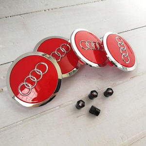 May include: Set of four red and silver Audi wheel center caps with the Audi logo, plus four black valve stem caps with the Audi logo. The caps are arranged on a white wooden surface.