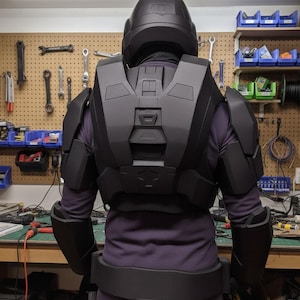 May include: Black cosplay armour set, including a helmet, chest piece, shoulder pads, and belt. The armour is worn over a purple long-sleeve shirt and trousers. The background shows a workshop with tools and equipment.