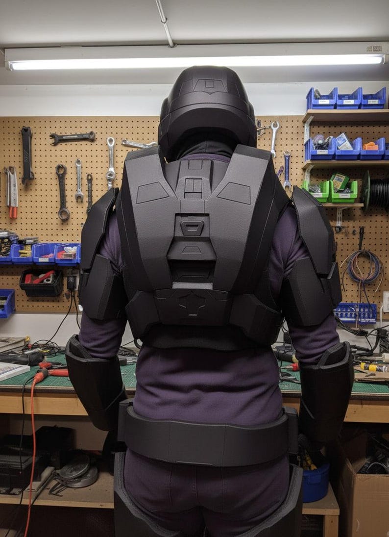 May include: Black cosplay armour set, including a helmet, chest piece, shoulder pads, and belt. The armour is worn over a purple long-sleeve shirt and trousers. The background shows a workshop with tools and equipment.