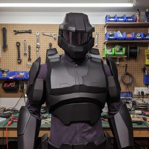 May include: A black and grey futuristic armour suit, including a helmet with a visor, chest and shoulder armour, and arm and leg guards. The suit is worn over a purple jumpsuit. The background shows a workshop with tools.