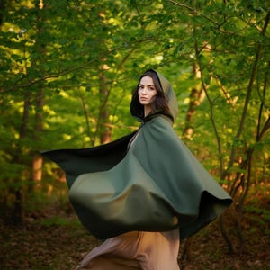 Pode incluir: Uma capa com capuz verde escuro com um design fluido. A capa é feita de um material macio e é usada ao ar livre em um ambiente florestal. A capa tem um capuz e cobre os ombros.
