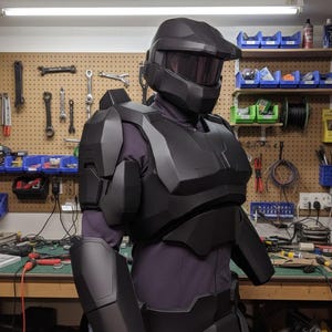 May include: A black futuristic armour set, comprising a helmet with a visor, chest and shoulder pieces, and arm guards. The armour is worn over a dark purple garment. The background shows a workshop with tools and equipment.
