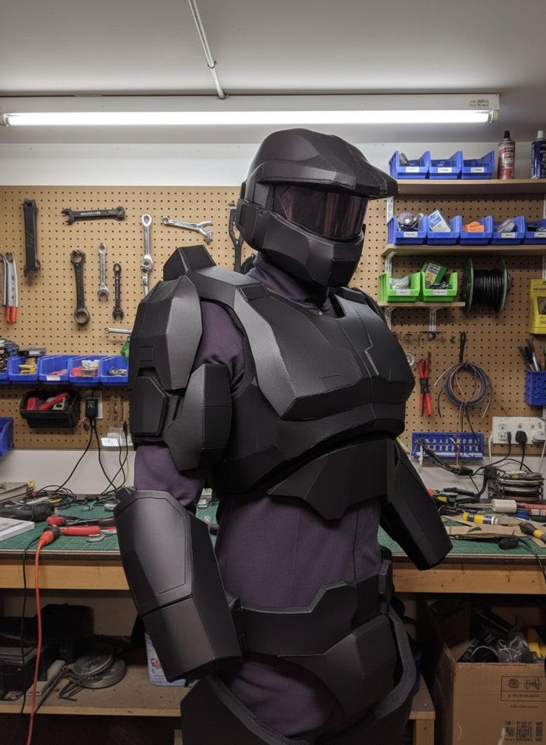 May include: A black futuristic armour set, comprising a helmet with a visor, chest and shoulder pieces, and arm guards. The armour is worn over a dark purple garment. The background shows a workshop with tools and equipment.