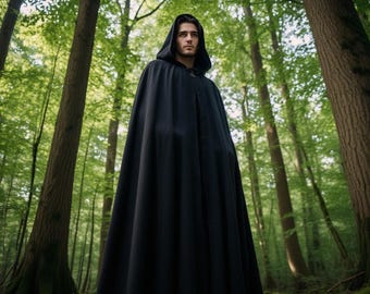 May include: A long, black hooded cloak with a clasp, set in a forest. The cloak drapes to the ground, with a deep hood. The background shows tall trees and green leaves, creating a mysterious atmosphere.