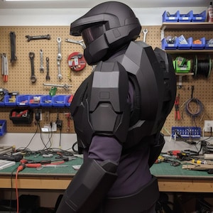 May include: A black futuristic armour set, including a helmet with a visor, chest and shoulder pieces, and arm guards. The armour is worn over a purple long-sleeved shirt. The background shows a workshop with tools and equipment.