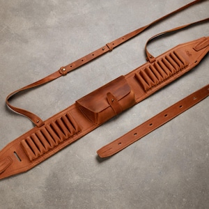 Può includere: Una cinghia per fucile in pelle color cuoio con due sezioni integrate per cartucce, una tasca centrale con chiusura a patta e una tracolla regolabile con fibbie in metallo. Il design presenta cuciture rinforzate e molteplici fori di regolazione.