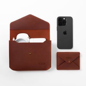 May include: A brown leather tech organizer with a matching wallet and a black smartphone. The organizer is open, revealing white accessories. The wallet and organizer have the name "JOHN" embossed on them.
