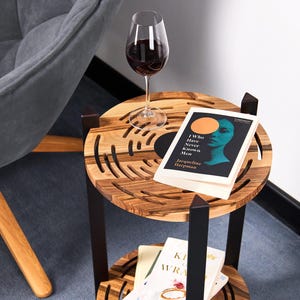 May include: A two-tiered wooden side table with a dark metal frame. The top tier holds a glass of red wine and a book titled "I Who Have Never Known Men." The bottom tier has books. The table is next to a gray armchair.