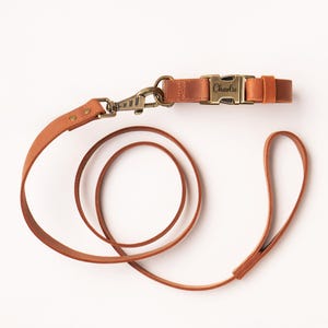 May include: A tan leather dog leash and collar set featuring antique brass hardware. The collar includes a side-release buckle engraved with the name Charlie. The leash has a swivel snap hook and a loop handle, connected by rivets to the strap.
