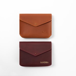 May include: Two rectangular leather pouches with envelope-style flaps and single metal snap closures. The top pouch is tan and embossed with the name Kevin. The bottom pouch is dark burgundy and embossed with the name SANDRA.