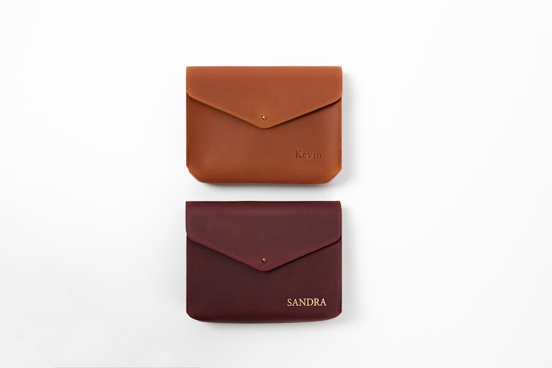 May include: Two rectangular leather pouches with envelope-style flaps and single metal snap closures. The top pouch is tan and embossed with the name Kevin. The bottom pouch is dark burgundy and embossed with the name SANDRA.