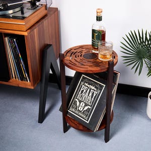 Puede incluir: Una mesa auxiliar de madera de dos niveles, con la parte superior en forma de disco de vinilo. Encima hay una botella de whisky y un vaso. La balda inferior contiene un álbum con la inscripción "PEAKY BLINDERS". Se ve un tocadiscos al fondo.