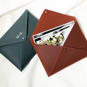 May include: Two leather envelope-style cases, one teal and one brown, are displayed open and closed. The teal case has the initials "M+R" and a snap closure. The brown case holds photos and a bouquet of flowers.