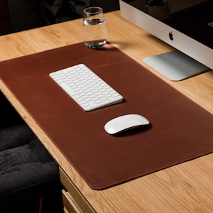 May include: A rectangular brown leather desk mat with rounded corners and a subtle embossed logo in the bottom corner. The mat provides a smooth surface for a white wireless keyboard and a white wireless computer mouse placed on top of it.