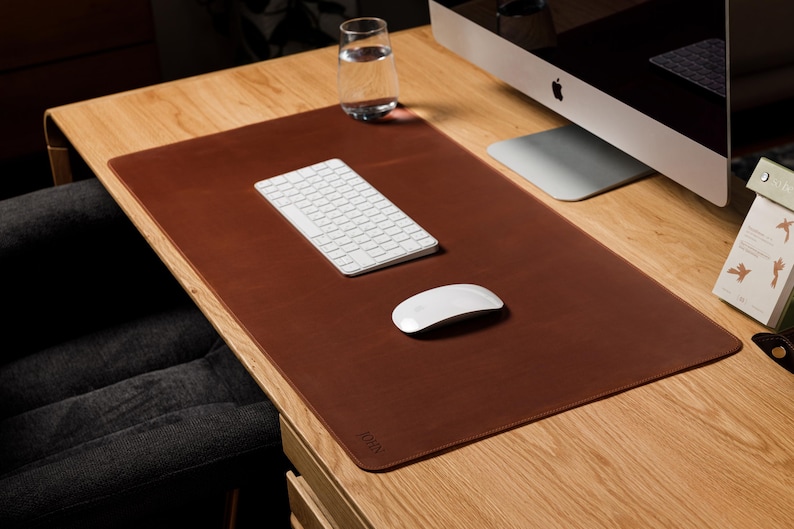 May include: A rectangular brown leather desk mat with rounded corners and a subtle embossed logo in the bottom corner. The mat provides a smooth surface for a white wireless keyboard and a white wireless computer mouse placed on top of it.