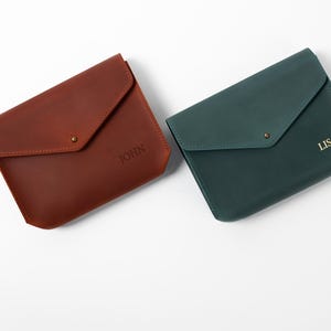 May include: Two leather envelope-style wallets. One is a reddish-brown color with the name "JOHN" embossed on the front. The other is a dark teal color with the name "LISA" embossed on the front. Each wallet has a flap closure with a small metal stud.