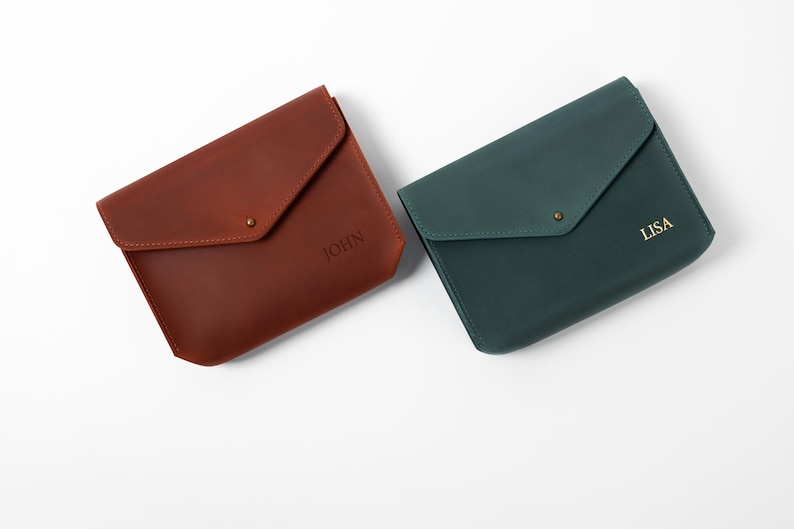 May include: Two leather envelope-style wallets. One is a reddish-brown color with the name "JOHN" embossed on the front. The other is a dark teal color with the name "LISA" embossed on the front. Each wallet has a flap closure with a small metal stud.
