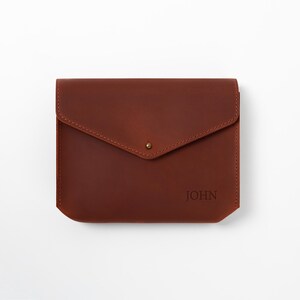 May include: A brown leather envelope-style case with a flap closure and a small gold-colored stud. The word "JOHN" is embossed on the lower right corner. The case is rectangular with rounded corners.