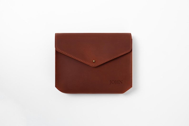 May include: A brown leather envelope-style case with a flap closure and a small gold-colored stud. The word "JOHN" is embossed on the lower right corner. The case is rectangular with rounded corners.