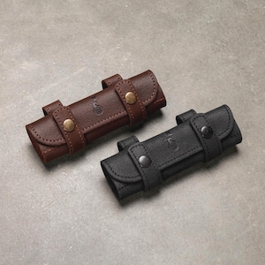 Peut inclure: Deux étuis rectangulaires en cuir, l'un marron foncé et l'autre noir, chacun doté de deux lanières avec boutons-pression métalliques et d'un petit logo gaufré sur le rabat avant. Les étuis présentent une fermeture à rabat et des coutures visibles.