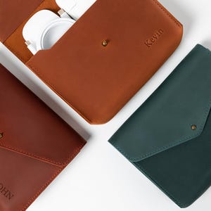 May include: Three leather cases in shades of brown and green are displayed on a white surface. One case is open, revealing white accessories. The cases are personalized with names: "John", "Kevin", and "Lisa". Each case has a flap closure with a gold-colored snap.
