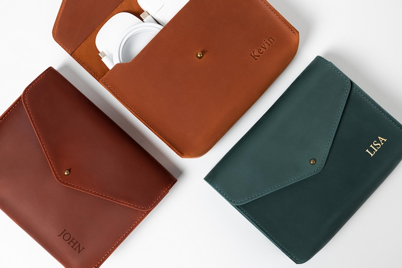 May include: Three leather cases in shades of brown and green are displayed on a white surface. One case is open, revealing white accessories. The cases are personalized with names: "John", "Kevin", and "Lisa". Each case has a flap closure with a gold-colored snap.