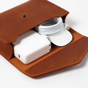 May include: A brown leather tech organizer with the name "Kevin" embossed on the flap. The open case reveals a white power adapter, a white charging cable, and a white mouse. The case is designed to hold tech accessories.