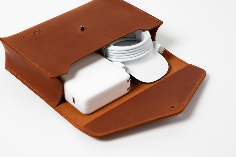 May include: A brown leather tech organizer with the name "Kevin" embossed on the flap. The open case reveals a white power adapter, a white charging cable, and a white mouse. The case is designed to hold tech accessories.