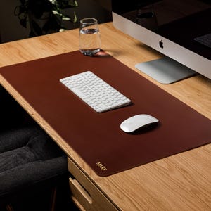 May include: A rectangular dark brown desk mat with rounded corners, featuring the name MARY embossed in gold lettering on the bottom left corner. The mat is placed on a wooden desk surface, providing a smooth area for a white keyboard and mouse.