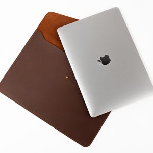 Könnte beinhalten: Ein silberner Laptop mit schwarzem Apple-Logo liegt neben einer braunen Laptop-Hülle aus Leder. Die Hülle hat eine hellbraune Innenseite und einen goldfarbenen Knopfverschluss. Die Artikel befinden sich auf weißem Hintergrund.