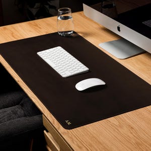 May include: A rectangular dark brown desk mat with rounded corners and the gold embossed initials A.M. in the bottom left corner. The mat provides a smooth surface for a white wireless keyboard and a white wireless computer mouse.