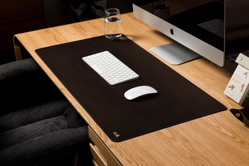 May include: A rectangular dark brown desk mat with rounded corners and the gold embossed initials A.M. in the bottom left corner. The mat provides a smooth surface for a white wireless keyboard and a white wireless computer mouse.