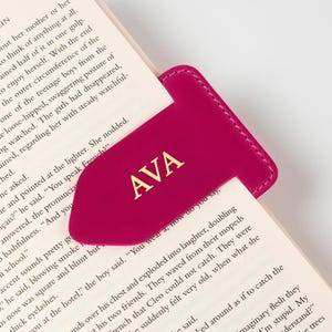 Könnte beinhalten: Ein magenta-farbener Leder-Lesezeichen mit den Initialen "AVA" in goldfarbenen Buchstaben. Das Lesezeichen ist in ein Buch eingelegt und unterstreicht seine Verwendung als Lesezubehör. Das Design des Lesezeichens ist schlicht und elegant.
