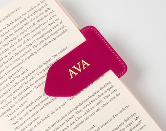 May include: A magenta leather bookmark with the initials "AVA" in gold lettering. The bookmark is inserted into a book, highlighting its use as a reading accessory. The bookmark's design is simple and elegant.