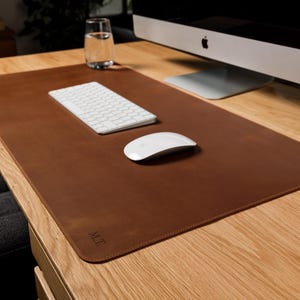May include: A rectangular brown leather desk mat with rounded corners and stitched edges. The mat features the embossed initials M.T. in the bottom left corner. It is placed on a wooden desk surface under a white computer keyboard and mouse.