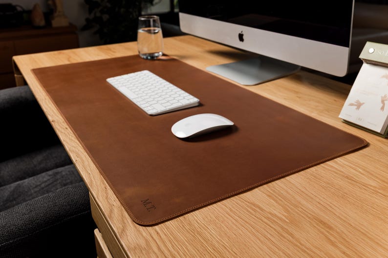May include: A rectangular brown leather desk mat with rounded corners and stitched edges. The mat features the embossed initials M.T. in the bottom left corner. It is placed on a wooden desk surface under a white computer keyboard and mouse.