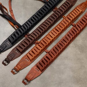 Könnte beinhalten: Vier Gewehrriemen aus Leder, diagonal angeordnet, mit integrierten Patronenschlaufen. Die Auswahl umfasst einen schwarzen Riemen und drei Brauntöne, jeweils ausgestattet mit einer Metallschnalle und verstellbaren Riemenelementen zur Befestigung.