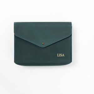 May include: A dark teal leather clutch with a flap closure and a gold-colored snap. The word "LISA" is printed in gold on the lower right. The clutch is rectangular and appears to be made of smooth leather.
