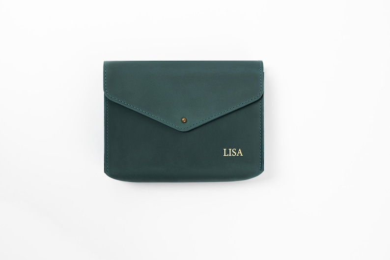 May include: A dark teal leather clutch with a flap closure and a gold-colored snap. The word "LISA" is printed in gold on the lower right. The clutch is rectangular and appears to be made of smooth leather.