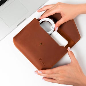 May include: A brown leather envelope-style pouch featuring a metal snap closure and the name Kevin embossed on the front. The pouch is open, revealing a white circular charging cable and a white power adapter stored inside the main compartment.