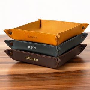 Puede incluir: Tres bandejas de cuero personalizadas apiladas sobre una superficie de madera. La bandeja superior es de color amarillo mostaza, la del medio es verde azulado oscuro y la inferior es marrón. Cada bandeja tiene un nombre grabado: "Lucia", "John" y "William".