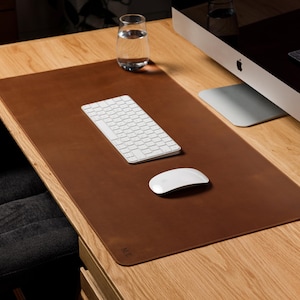 Op de afbeelding: Een rechthoekige bureaulegger van bruin leer met afgeronde hoeken en een glad oppervlak. De mat ligt op een houten bureau en dient als ondergrond voor een wit draadloos toetsenbord en een witte draadloze computermuis.