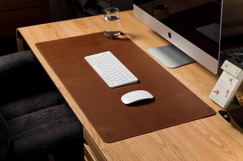 May include: A rectangular brown leather desk mat with rounded corners, featuring a smooth surface texture. The mat is positioned on a wooden desk surface, providing a base for a white wireless keyboard and a white wireless computer mouse.