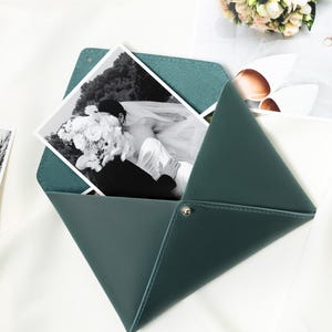 May include: A dark teal leather photo holder with a black and white wedding photo inside. The holder is open, revealing the photo, and is secured with a silver snap. Other wedding photos and a bouquet are in the background.