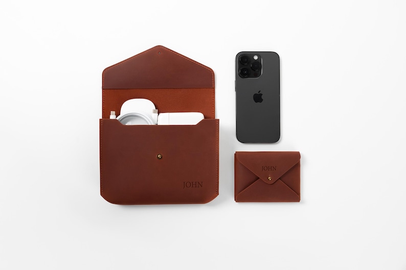 May include: A brown leather tech organizer with a matching wallet and a black smartphone. The organizer is open, revealing white accessories. The wallet and organizer have the name "JOHN" embossed on them.