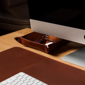 May include: A square brown leather valet tray with snap-button corners, containing a set of metal keys and a small white object. The tray is positioned on a wooden desk surface next to a computer monitor and a large brown leather desk mat.